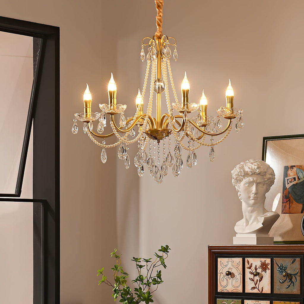 A Clérachic gold chandelier with candle-style bulbs lit, hung in a warm room beside a white plaster bust and a painted wooden cabinet.