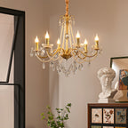 A Clérachic gold chandelier with candle-style bulbs lit, hung in a warm room beside a white plaster bust and a painted wooden cabinet.