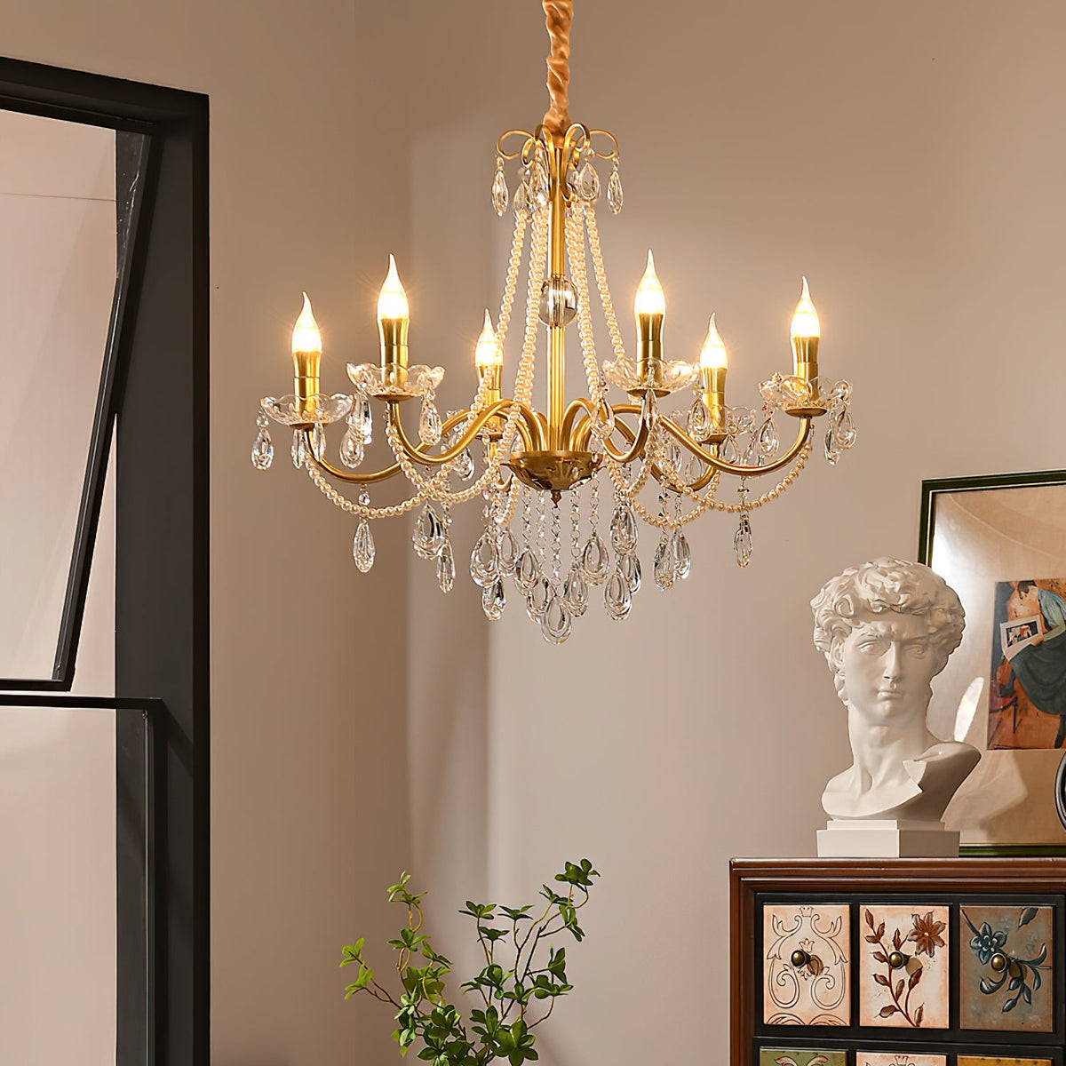 A Clérachic gold chandelier with candle-style bulbs lit, hung in a warm room beside a white plaster bust and a painted wooden cabinet.