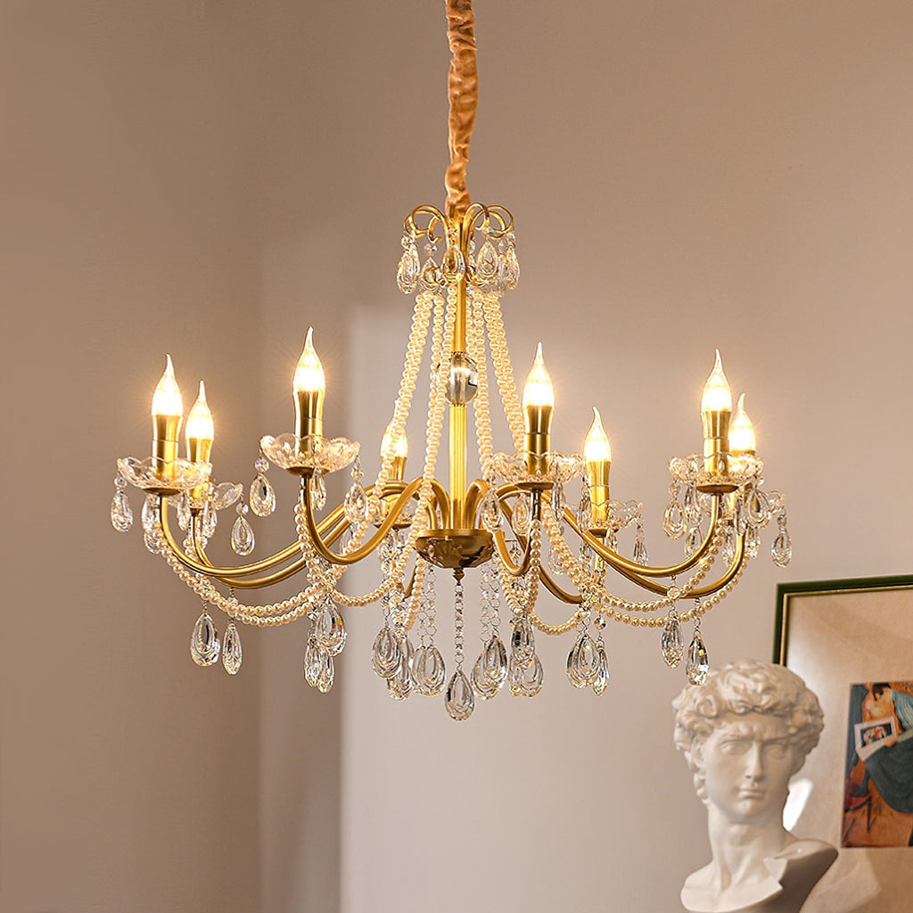 Lit candle-style bulbs on the curving gold arms of this Clérachic chandelier, with crystal drops below and a framed print at the side.