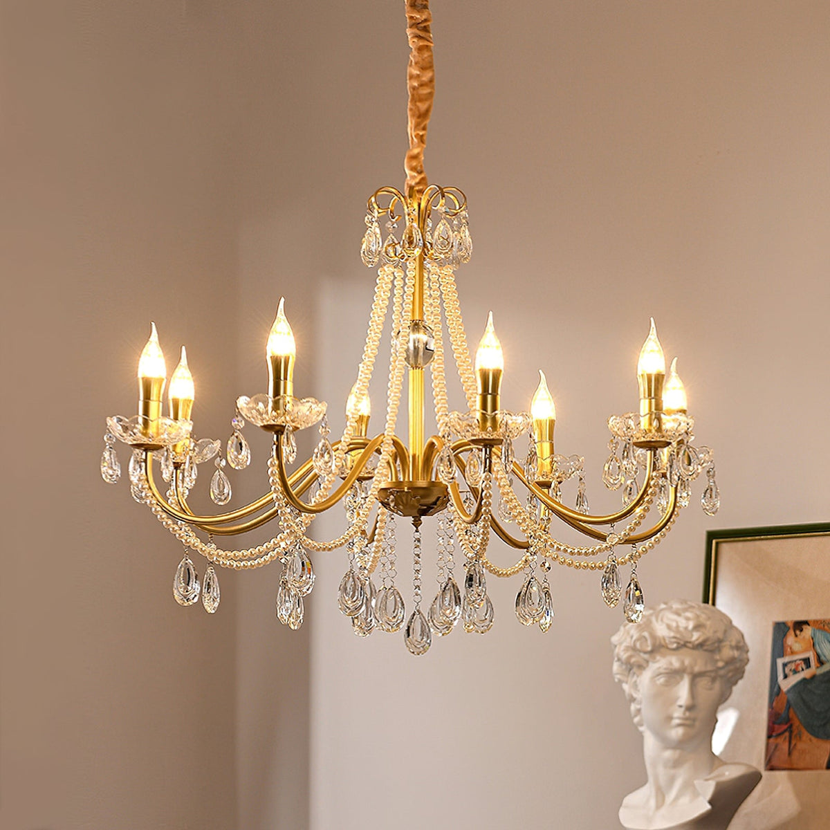 Lit candle-style bulbs on the curving gold arms of this Clérachic chandelier, with crystal drops below and a framed print at the side.