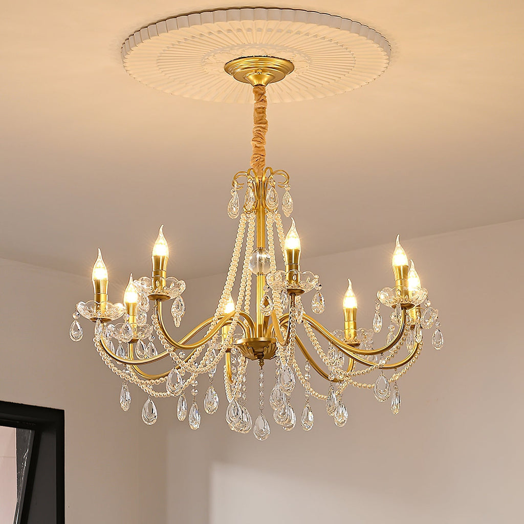 A Clérachic gold crystal chandelier hung from a ceiling rose with a gold canopy, in a cream room with a dark window frame.