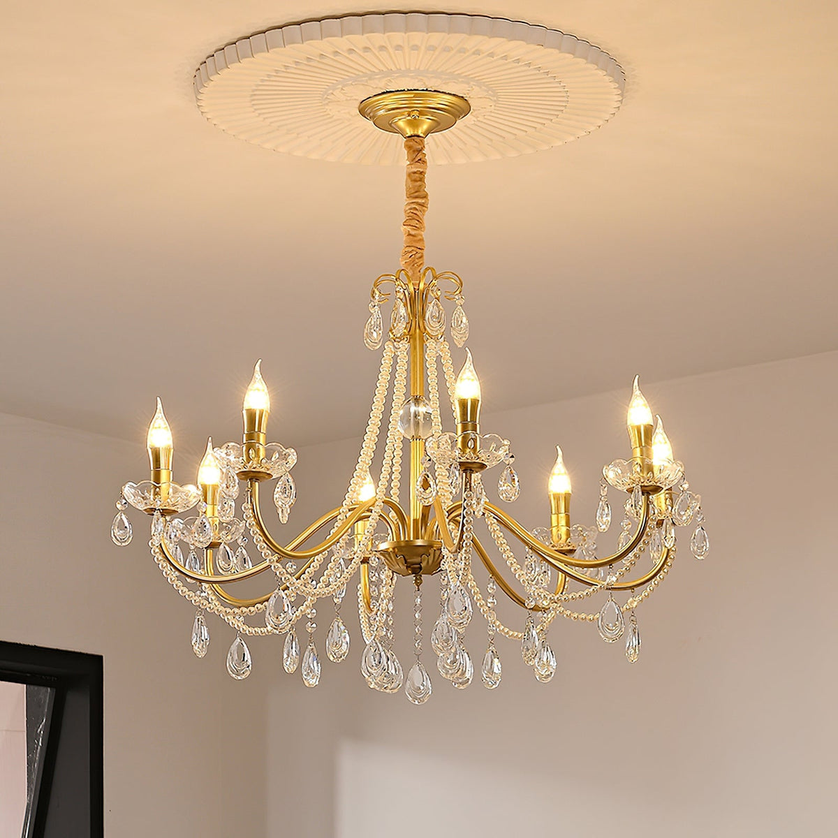 A Clérachic gold crystal chandelier hung from a ceiling rose with a gold canopy, in a cream room with a dark window frame.