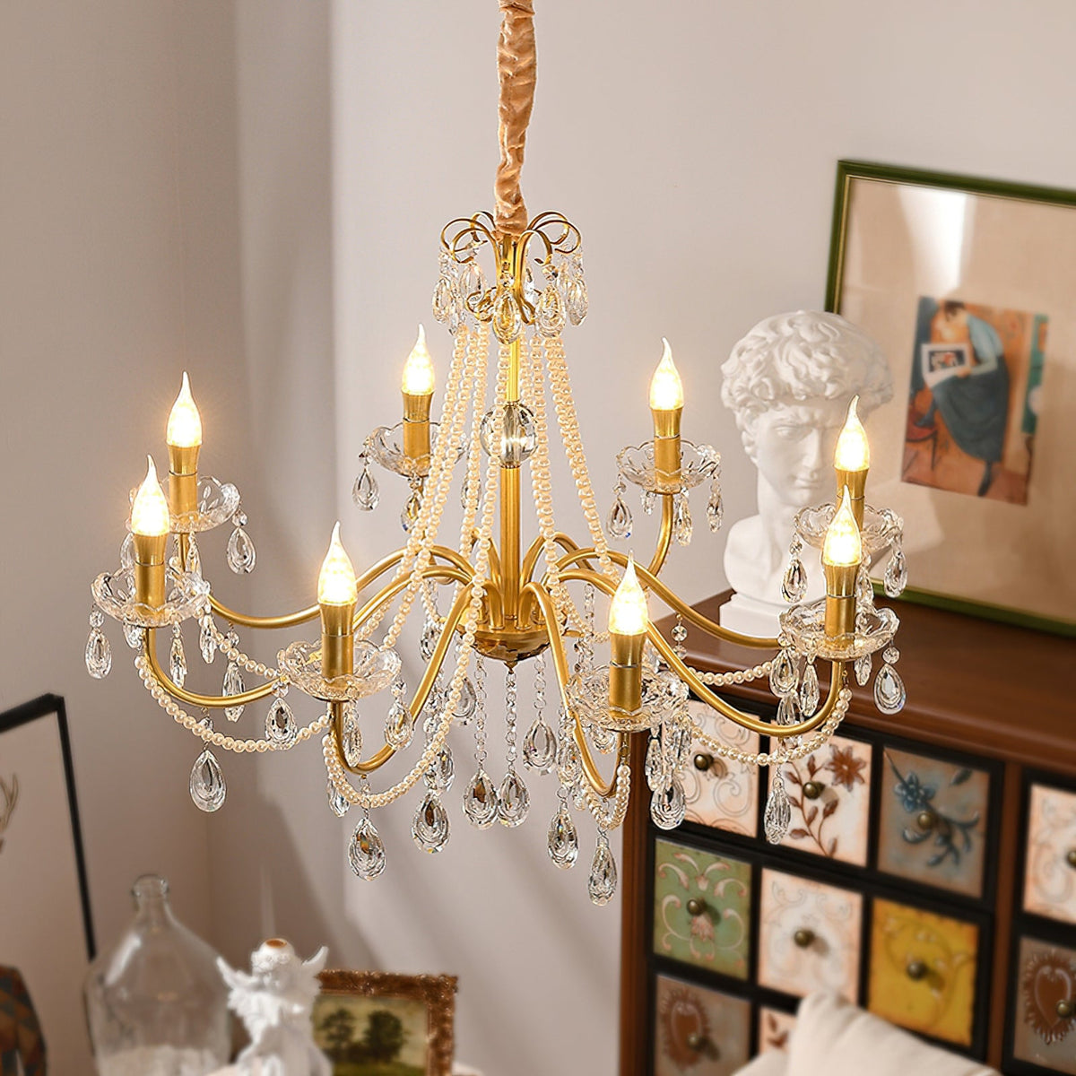 A Clérachic gold chandelier lit warmly above a painted wooden drawer unit, with a white plaster bust and a framed print beside it.