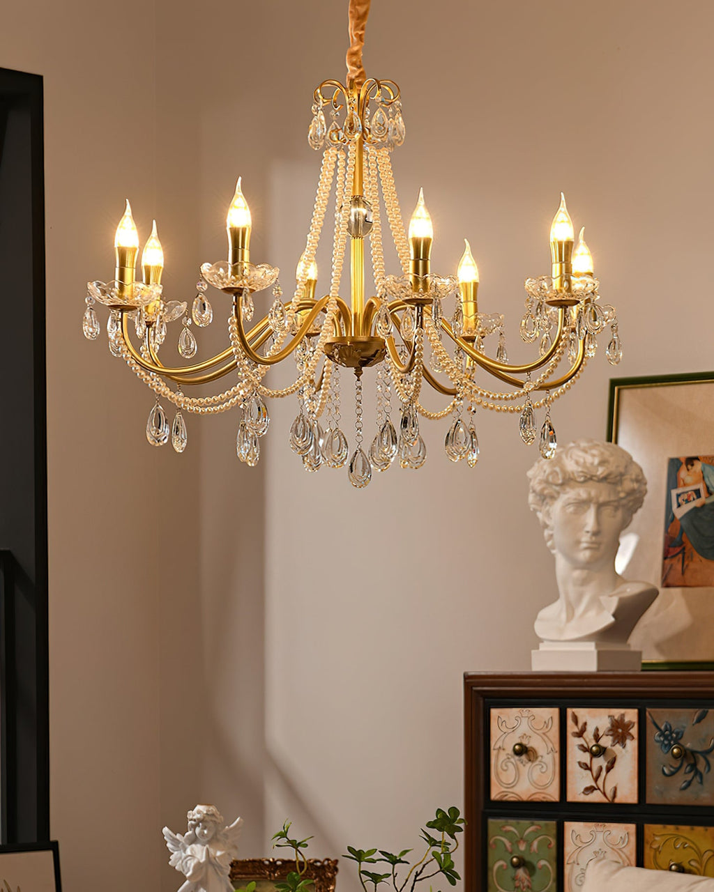 A Clérachic gold chandelier lit in a corner of a warm room, with a painted drawer cabinet and a framed print below its crystal drops.