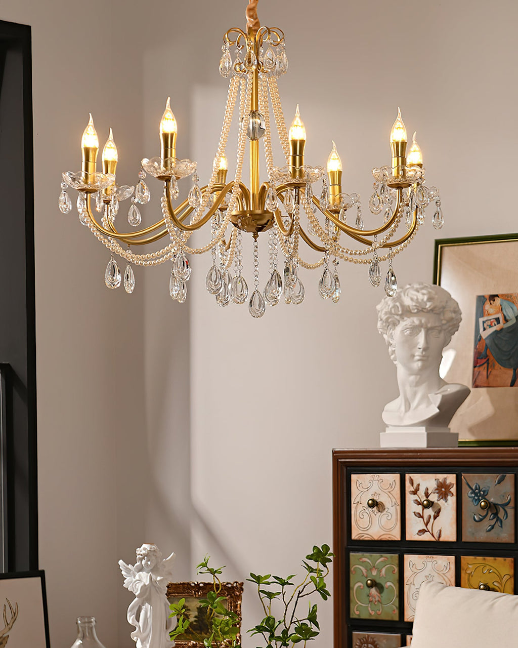 A Clérachic gold and crystal chandelier lit above a drawer cabinet with a plaster bust, an angel figurine and a green plant.