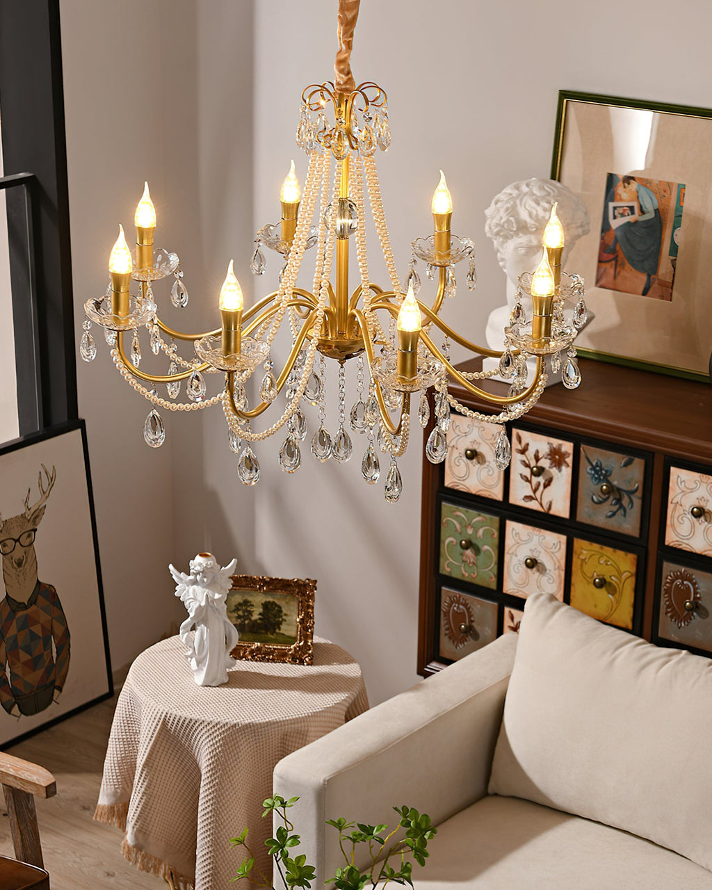 A Clérachic chandelier with lit candle bulbs and crystal drops above a beige sofa, a draped round table and a painted drawer cabinet.