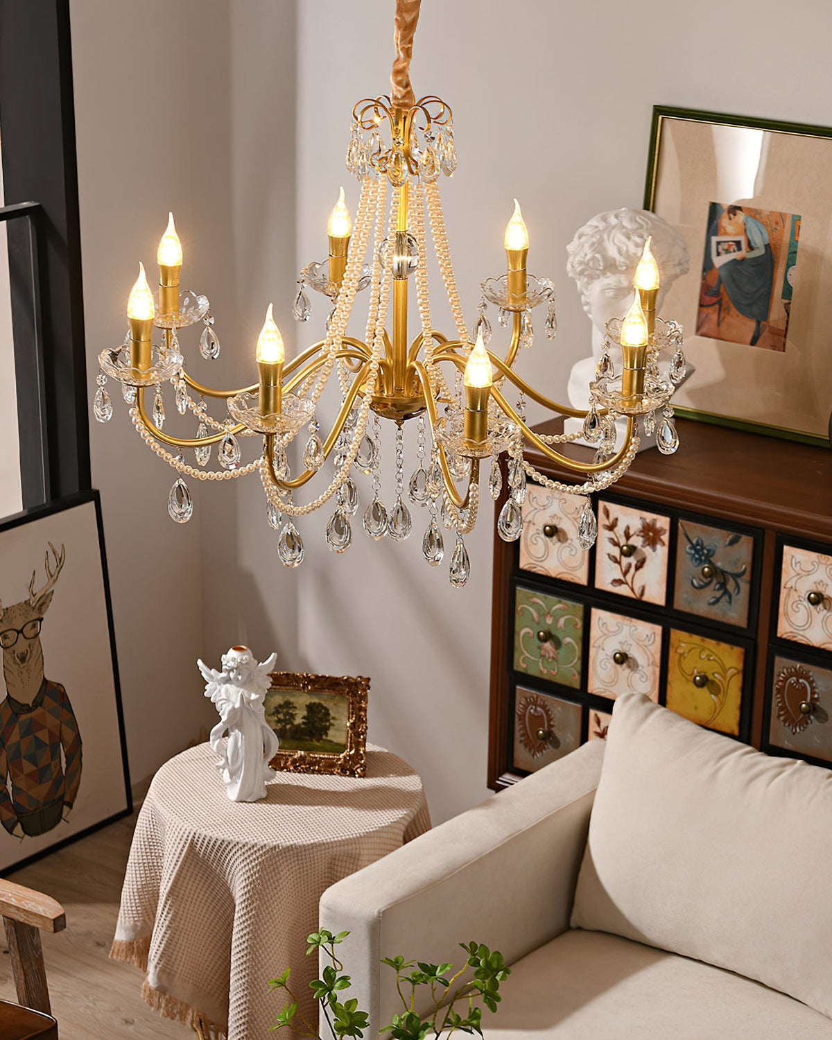 A Clérachic chandelier with lit candle bulbs and crystal drops above a beige sofa, a draped round table and a painted drawer cabinet.