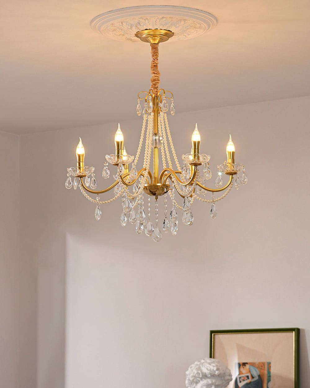 A Clérachic gold chandelier hung from a decorative ceiling rose, with lit candle bulbs and crystal drops in a pale room.