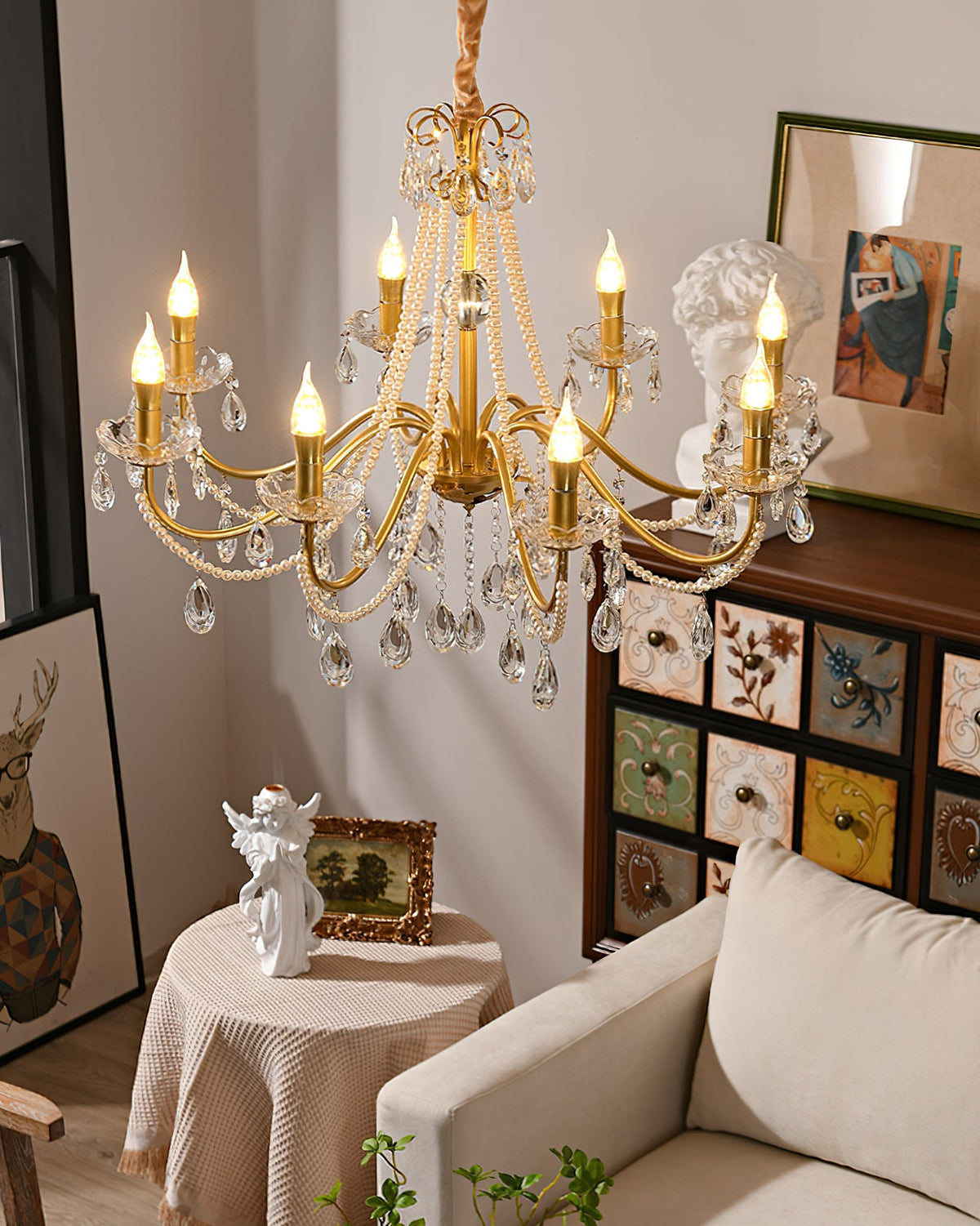 The same Clérachic chandelier lit over a draped round table, a beige sofa and a painted drawer cabinet in the corner of a room.