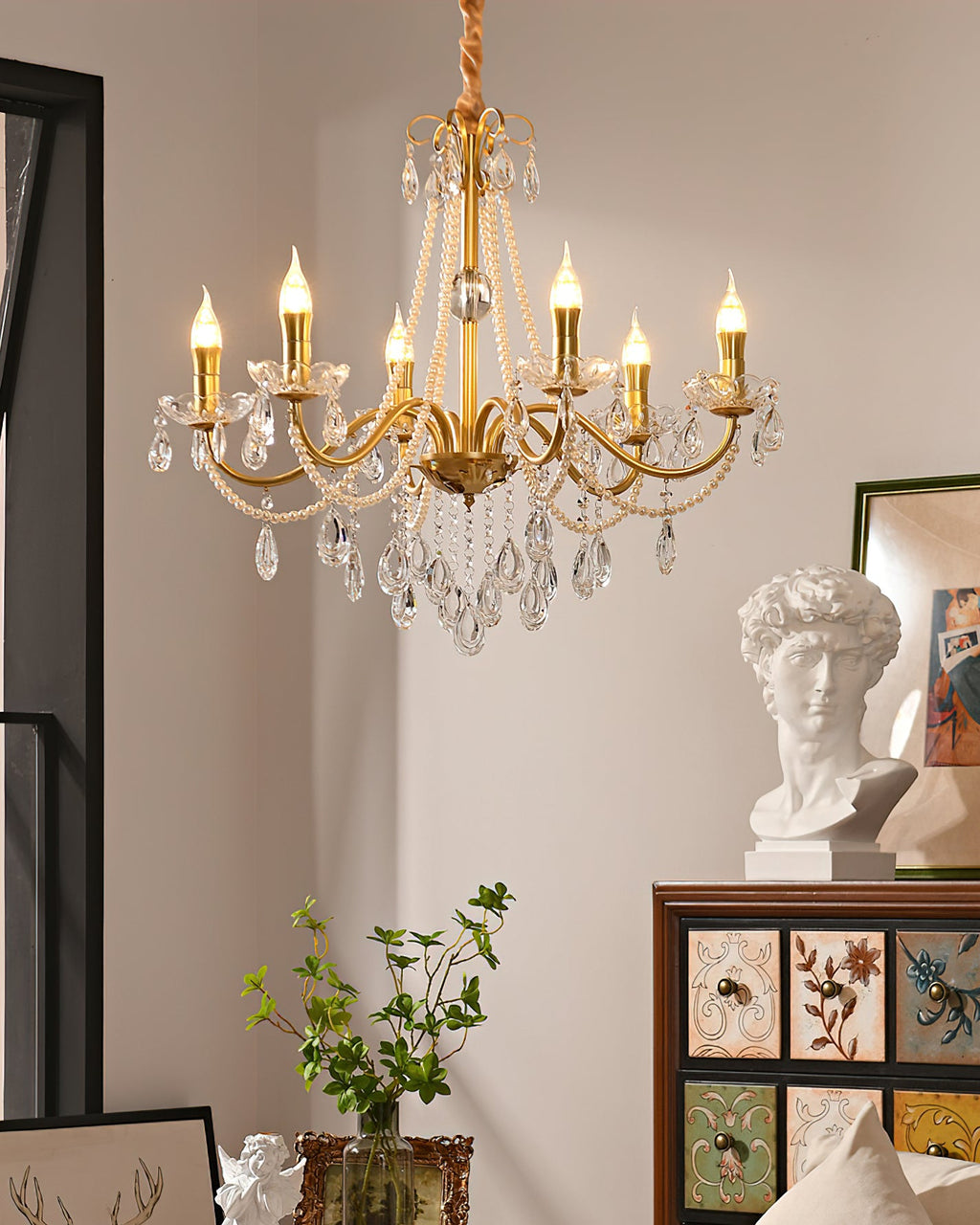 A Clérachic gold chandelier with lit candle bulbs and pearl strands, with a plaster bust and a leafy stem on a cabinet below.