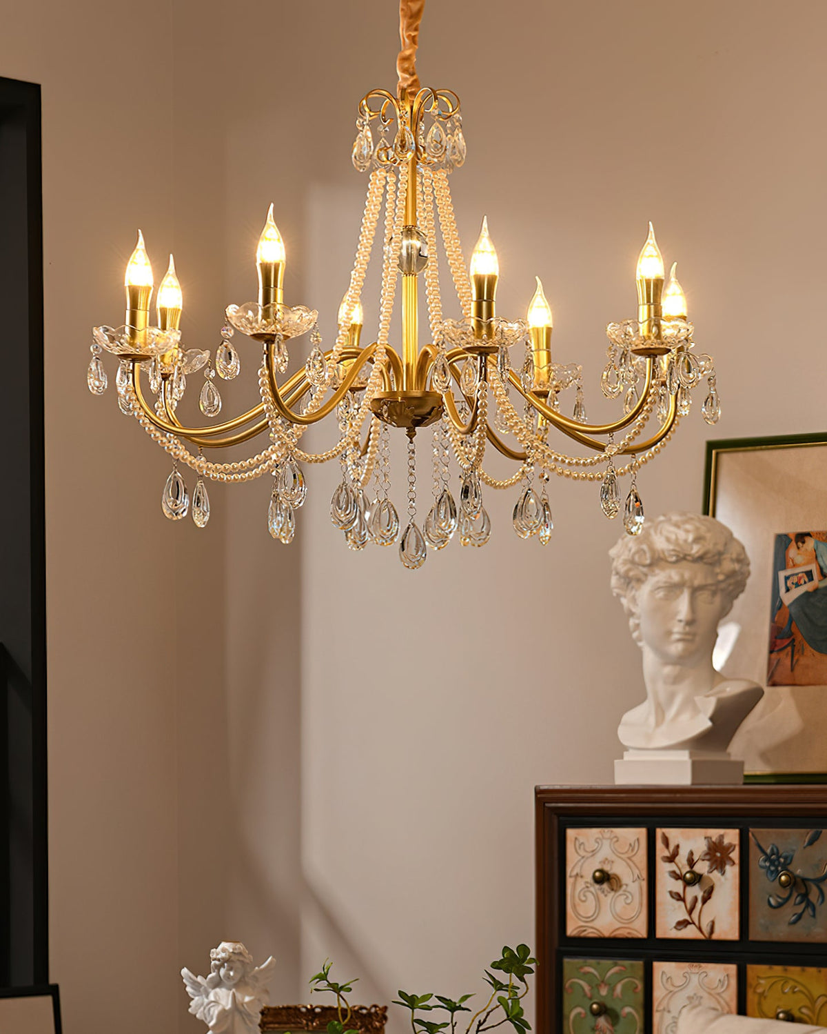 A Clérachic gold crystal chandelier glowing above a plaster bust, a framed print and a painted drawer cabinet in a warm room.
