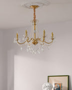 A Clérachic gold chandelier with soft candle-style bulbs, hung under a white plaster ceiling rose in a pale room.