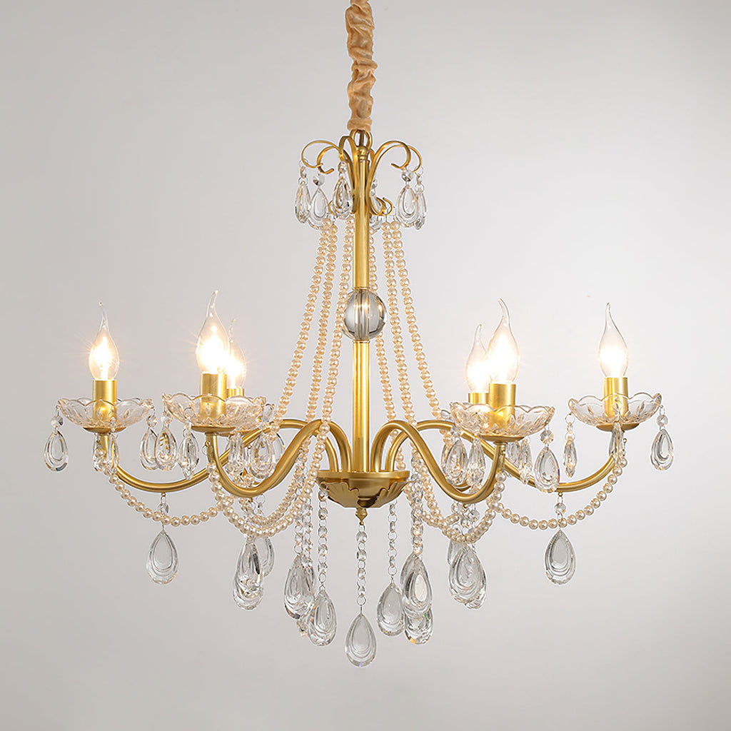 A Clérachic gold chandelier with lit candle-style bulbs, crystal drops and pearl strands on a soft grey backdrop.