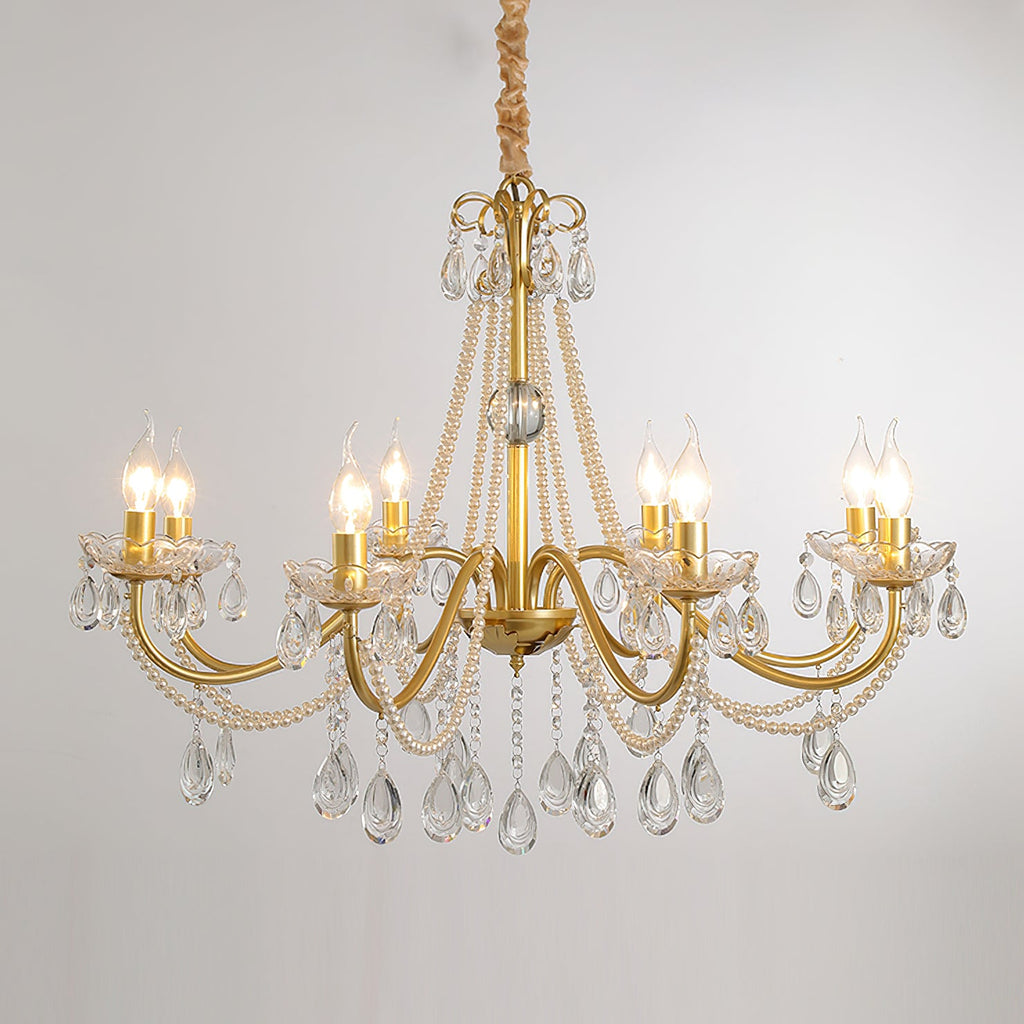 A Clérachic chandelier with lit flame-tip bulbs on gold arms, hung against a pale grey backdrop with crystal drops below.