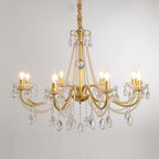 A Clérachic chandelier with lit flame-tip bulbs on gold arms, hung against a pale grey backdrop with crystal drops below.