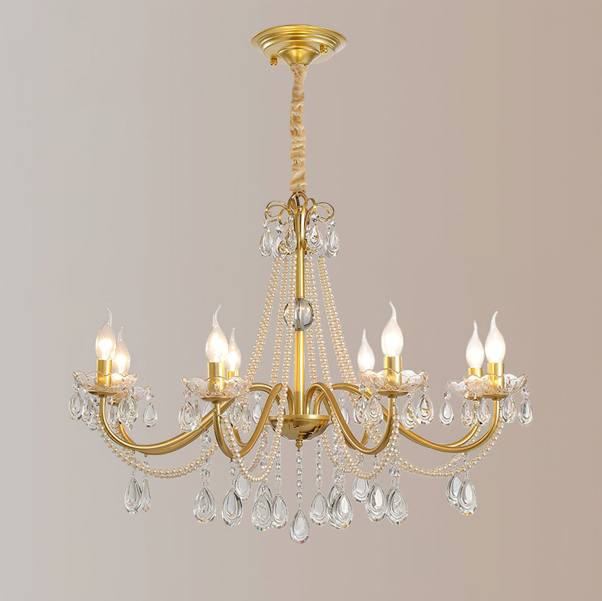 A Clérachic gold chandelier on a gold ceiling canopy and a wrapped chain, with candle bulbs lit against a blush-grey backdrop.