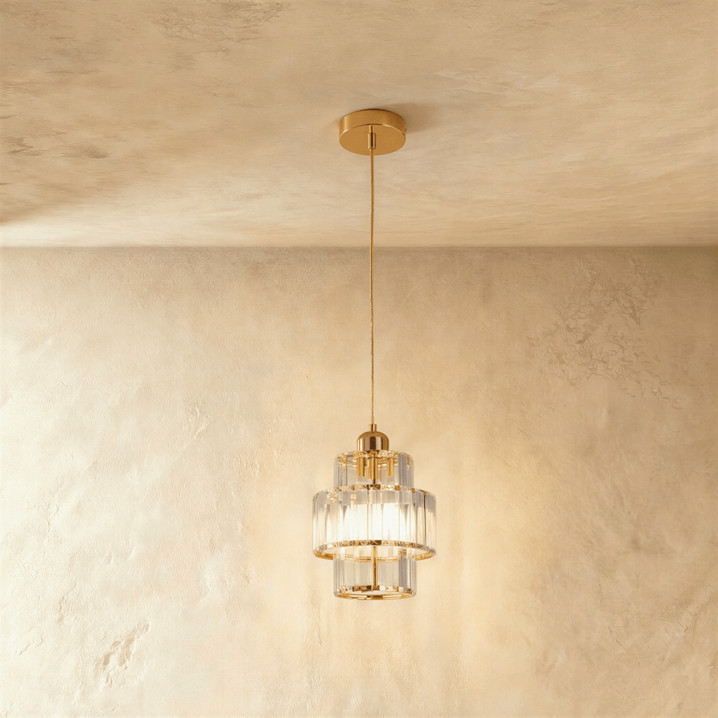 Clérachic tiered crystal pendant with a gold canopy and cord, lit and glowing against a limewash plaster wall.