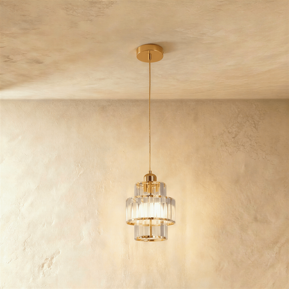 Clérachic tiered crystal pendant with a gold canopy and cord, lit and glowing against a limewash plaster wall.