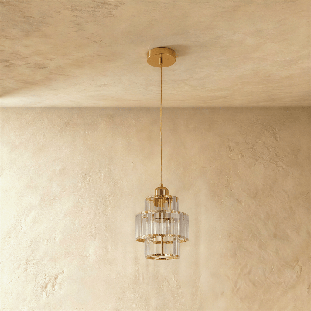 Clérachic three-tier crystal pendant on a gold cord and canopy, unlit, hung against a soft plaster wall.