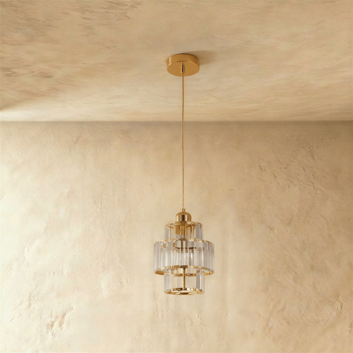 Clérachic three-tier crystal pendant on a gold cord and canopy, unlit, hung against a soft plaster wall.