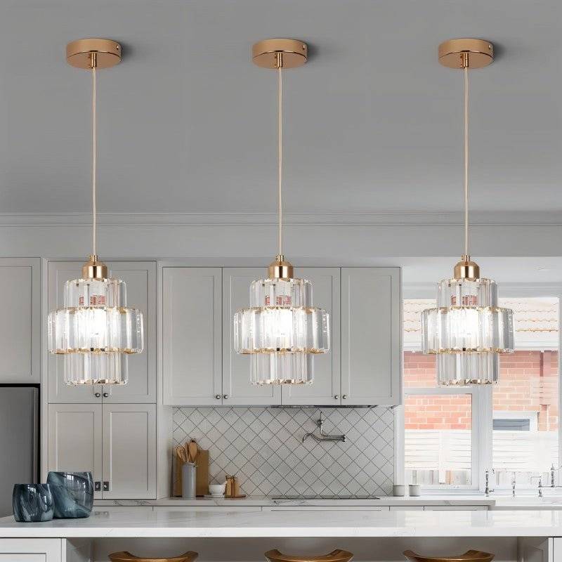 Three Clérachic gold-framed crystal pendants hung in a row above a white kitchen island with cabinets behind.