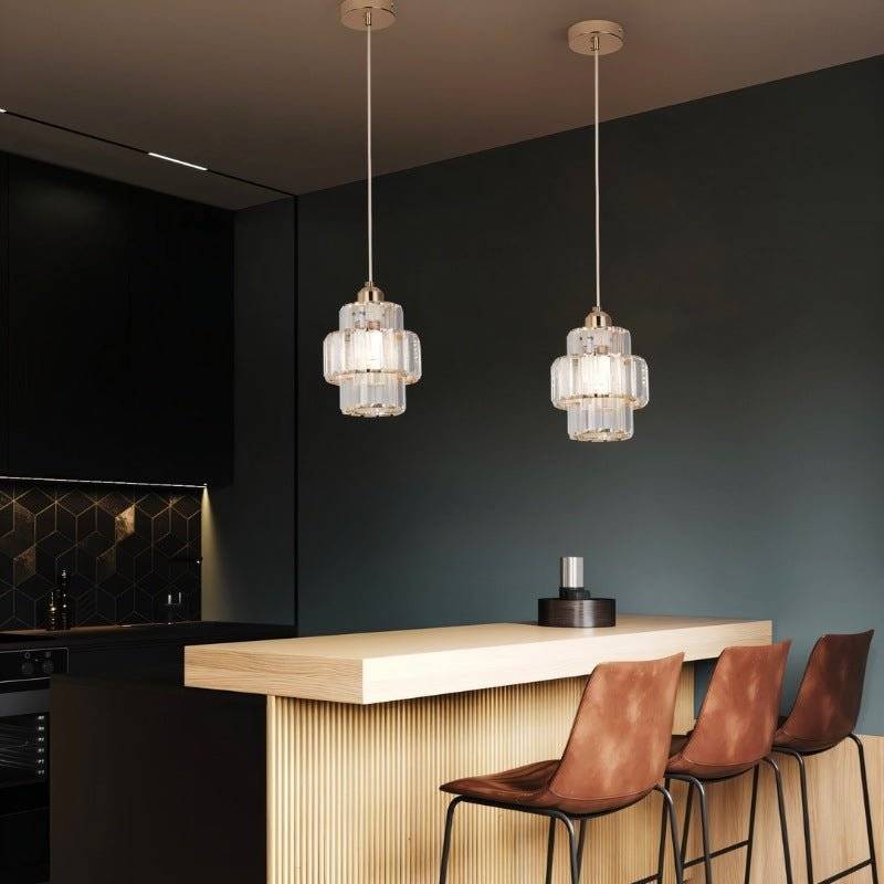 Two Clérachic gold crystal pendants hung above a pale wood breakfast bar in a dark green kitchen.