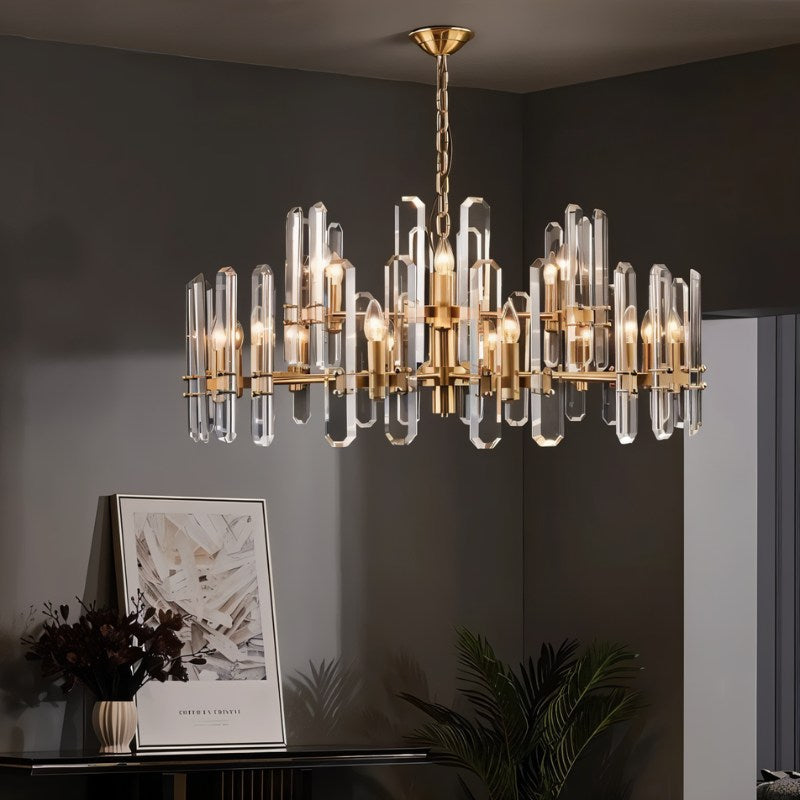 Clérachic round crystal-rod chandelier in brass with candle bulbs, hung in a grey room beside a framed print and a plant.
