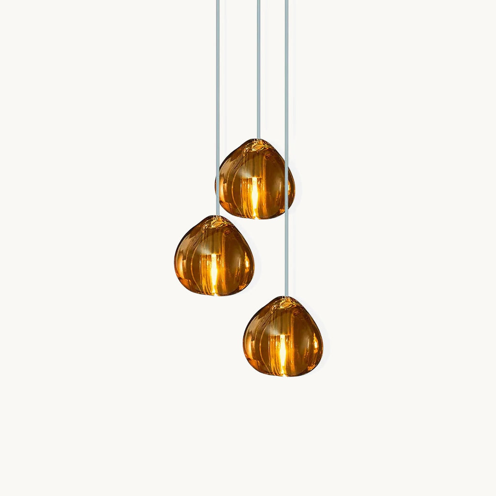 This Clérachic glass pendant on a white backdrop has three amber glass heads hanging at different heights on pale cords.