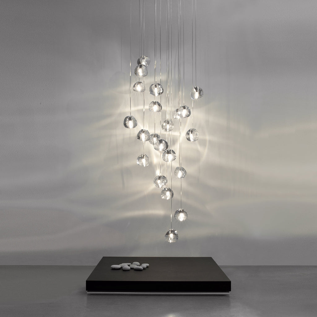A tall cluster of clear glass heads on this Clérachic pendant hangs from fine wires in a grey room, casting rippling light on the wall behind a low black plinth.