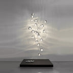 A tall cluster of clear glass heads on this Clérachic pendant hangs from fine wires in a grey room, casting rippling light on the wall behind a low black plinth.