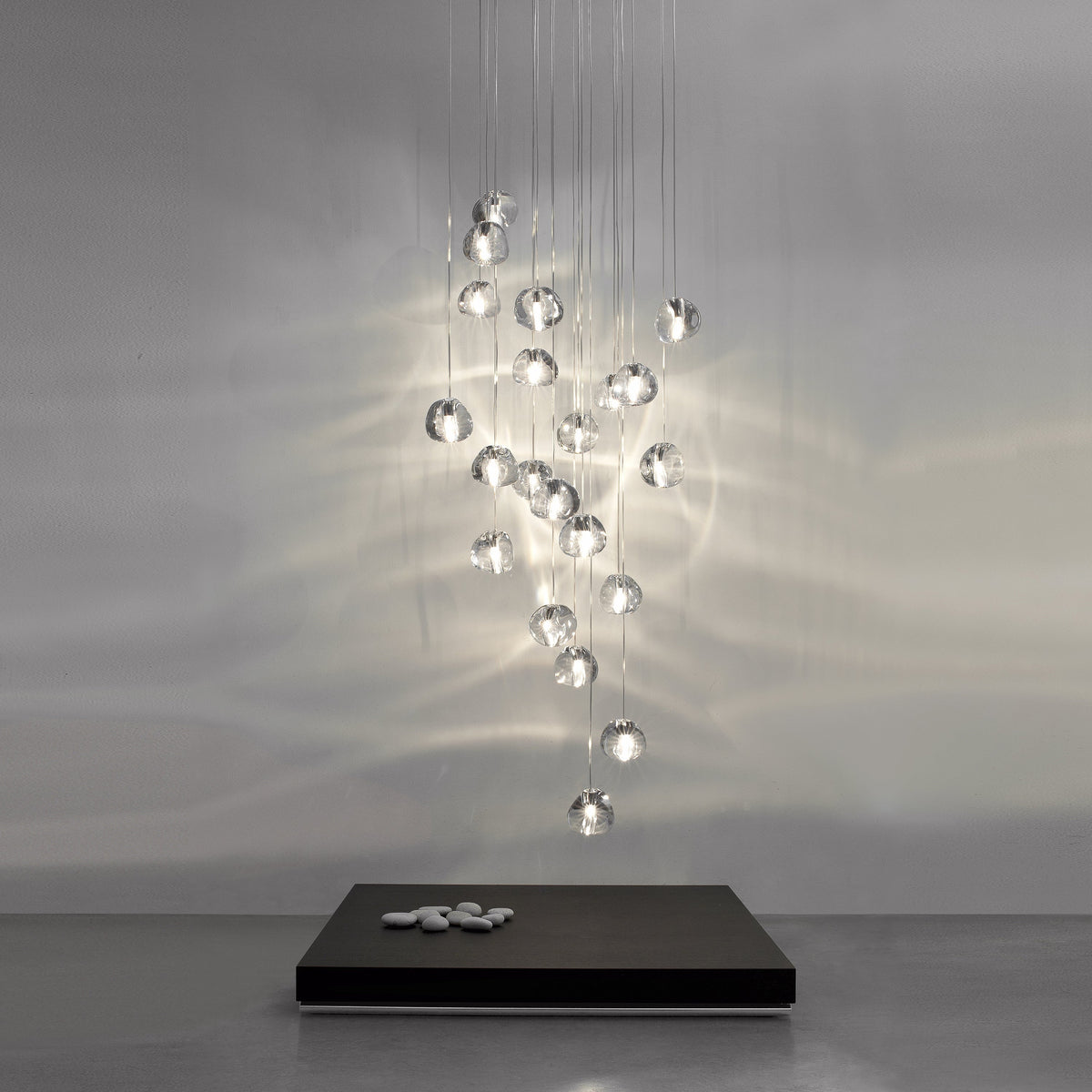 A tall cluster of clear glass heads on this Clérachic pendant hangs from fine wires in a grey room, casting rippling light on the wall behind a low black plinth.