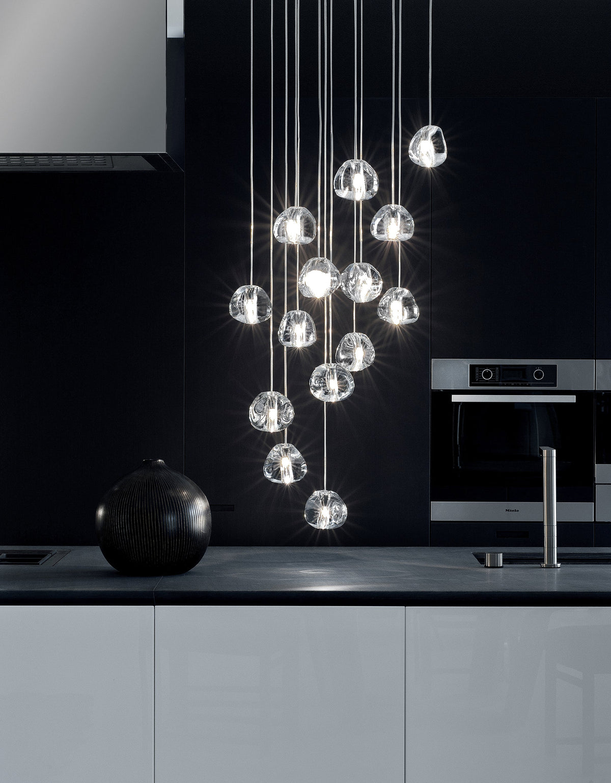 Clear glass heads of this Clérachic pendant sparkle in a black kitchen above a dark worktop, with a round black vase beside them.