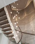 A Clérachic pendant cluster hangs through a spiral timber staircase, its clear heads glowing against a warm copper-toned wall.