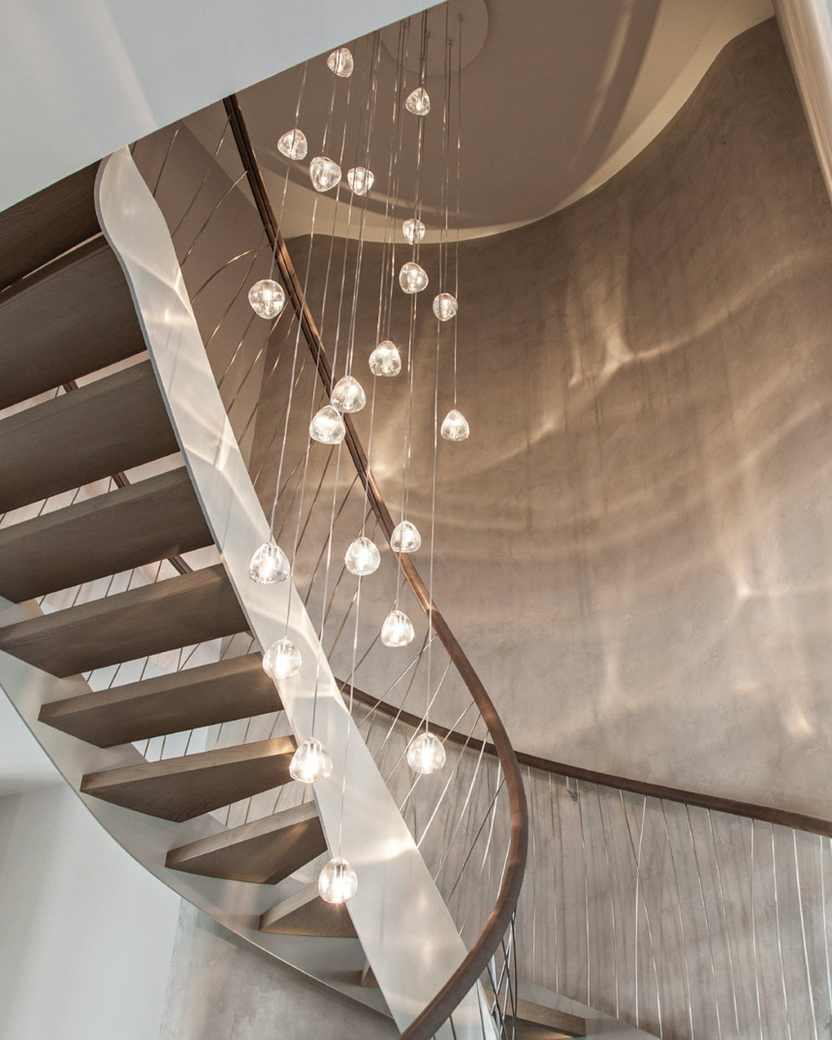 A Clérachic pendant cluster hangs through a spiral timber staircase, its clear heads glowing against a warm copper-toned wall.
