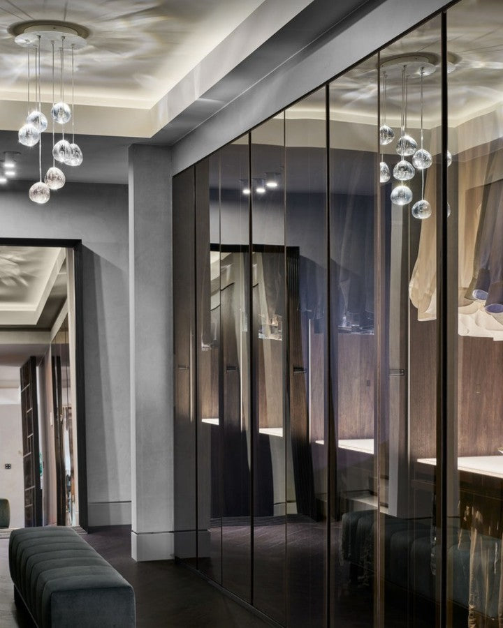 Clear glass heads of this Clérachic pendant hang in a dressing room with mirrored doors, beside a dark green bench.