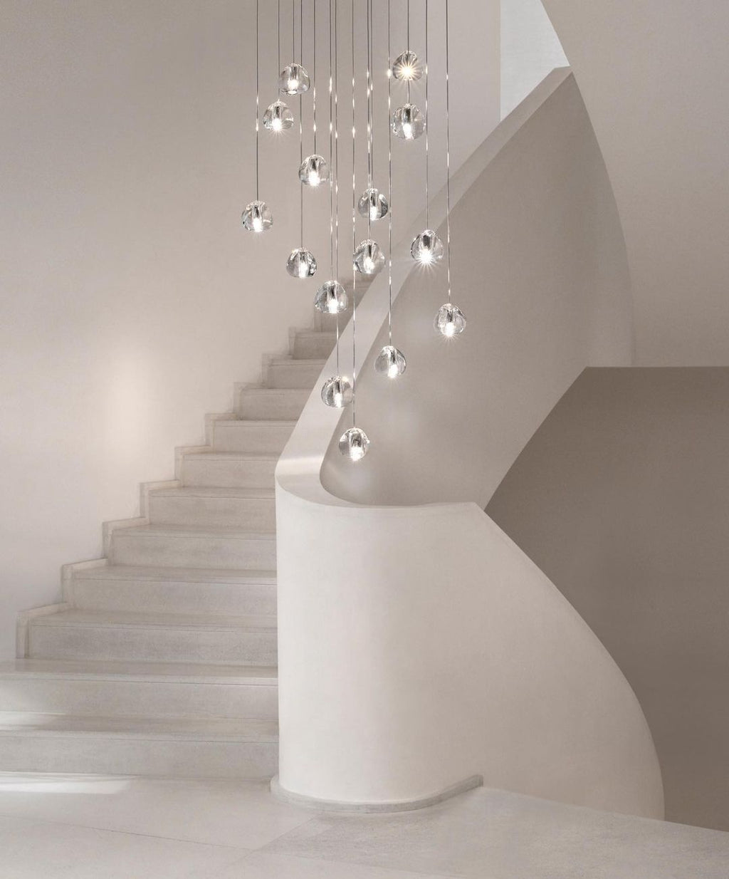 A Clérachic pendant cluster of clear glass heads hangs above a white curved staircase in a softly lit hall.