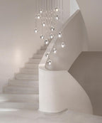 A Clérachic pendant cluster of clear glass heads hangs above a white curved staircase in a softly lit hall.