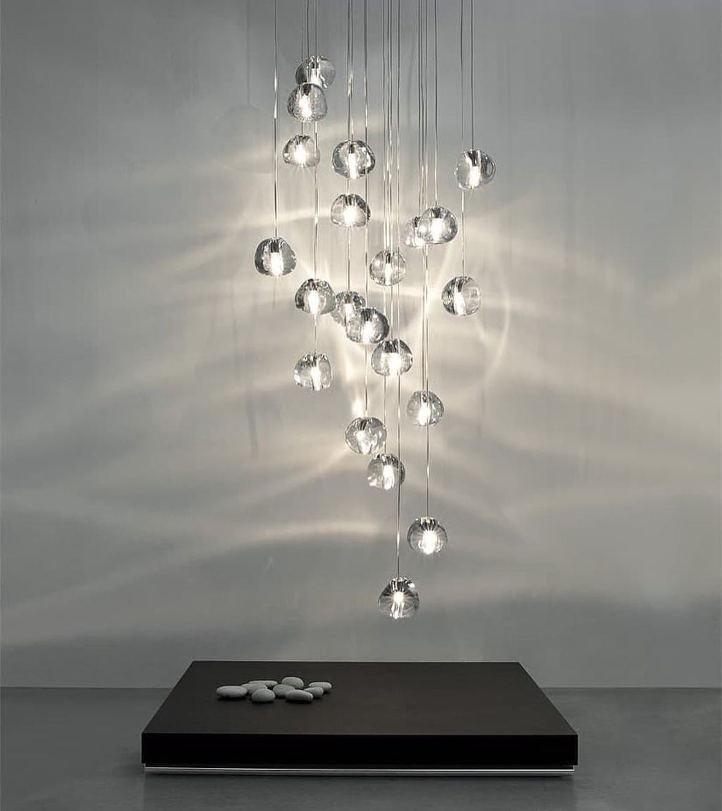 This Clérachic pendant hangs as a long cluster of clear glass heads, throwing soft ripples of light over a grey wall and a dark plinth.