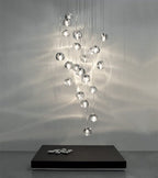 This Clérachic pendant hangs as a long cluster of clear glass heads, throwing soft ripples of light over a grey wall and a dark plinth.