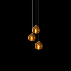 Three amber glass heads from this Clérachic pendant hang at staggered heights on pale cords against black.