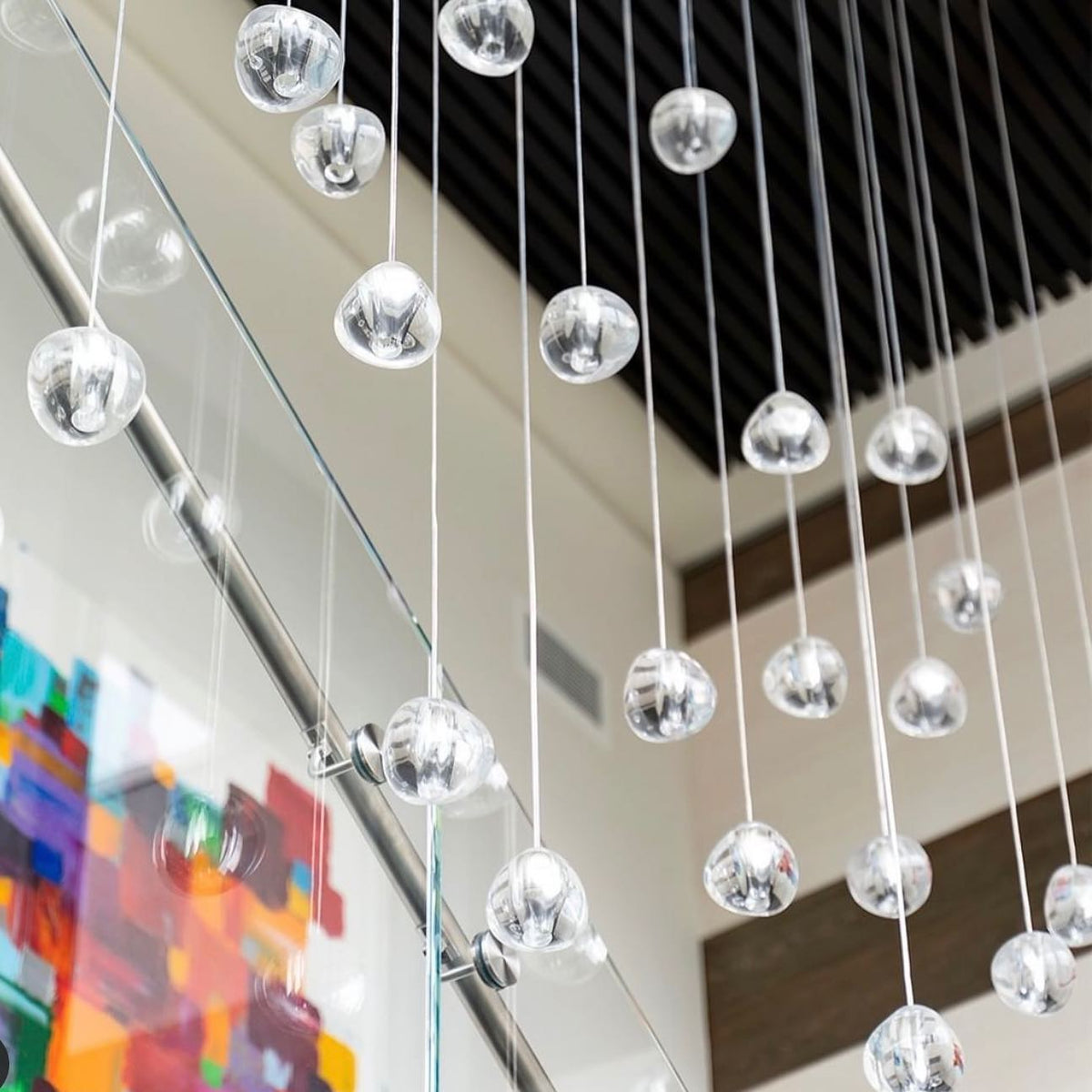 Close view of clear glass heads on long wires beside a glass balustrade, with a painting and a black slatted ceiling behind this Clérachic pendant.