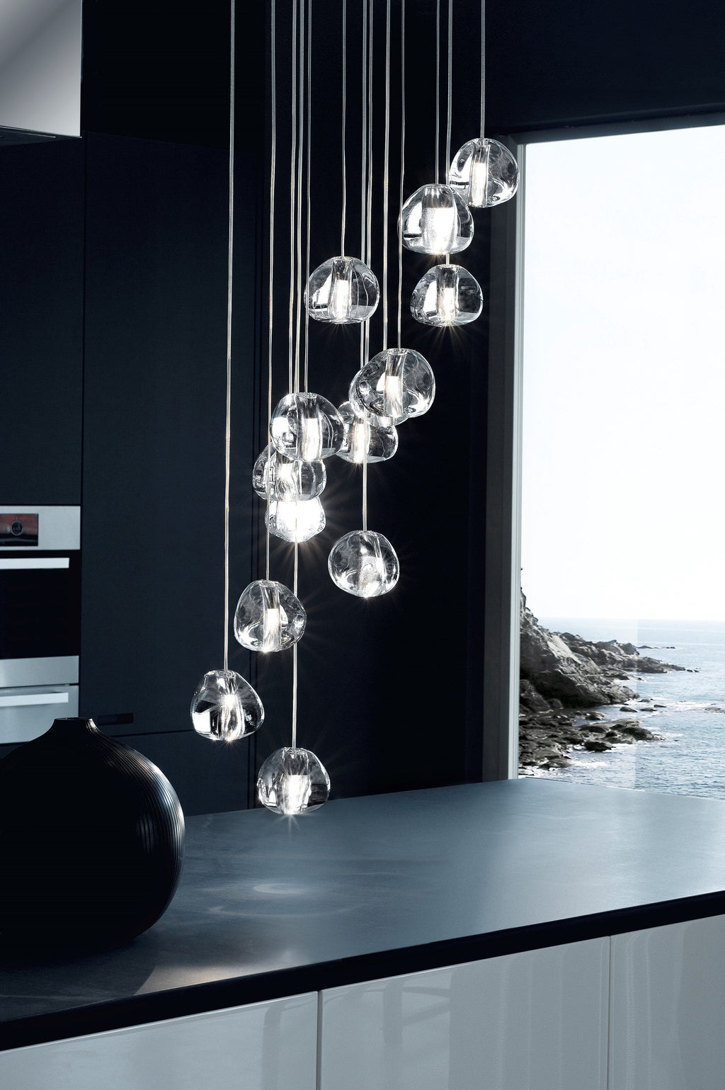 A tall, narrow view of this Clérachic pendant's clear glass heads above a black worktop, beside a window looking out to the sea.