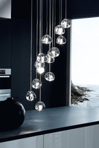 A tall, narrow view of this Clérachic pendant's clear glass heads above a black worktop, beside a window looking out to the sea.