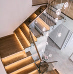 Looking down a timber staircase with lit treads and a glass balustrade, this Clérachic pendant's clear heads hang beside the flight.