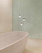 Three clear glass heads of this Clérachic pendant hang over a pale freestanding bath against a soft green wall.