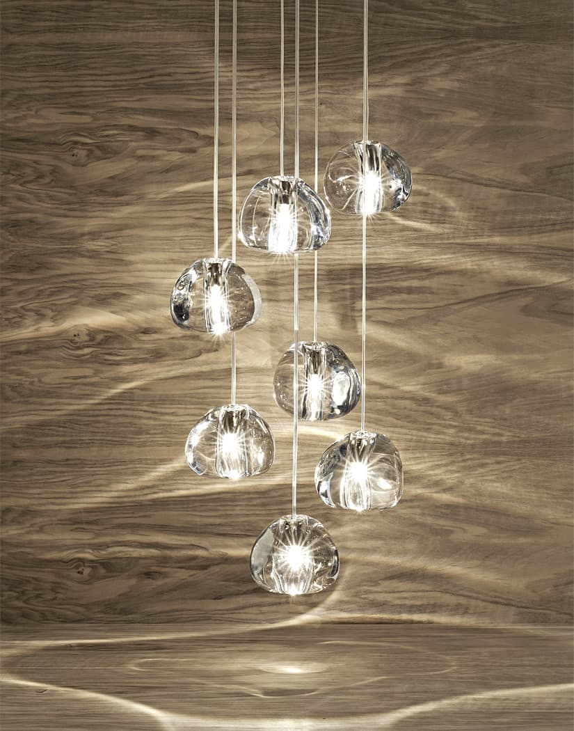 Seven clear glass heads of this Clérachic pendant hang in front of a driftwood-grain wall, which is marked with soft rippling reflections.