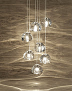 Seven clear glass heads of this Clérachic pendant hang in front of a driftwood-grain wall, which is marked with soft rippling reflections.