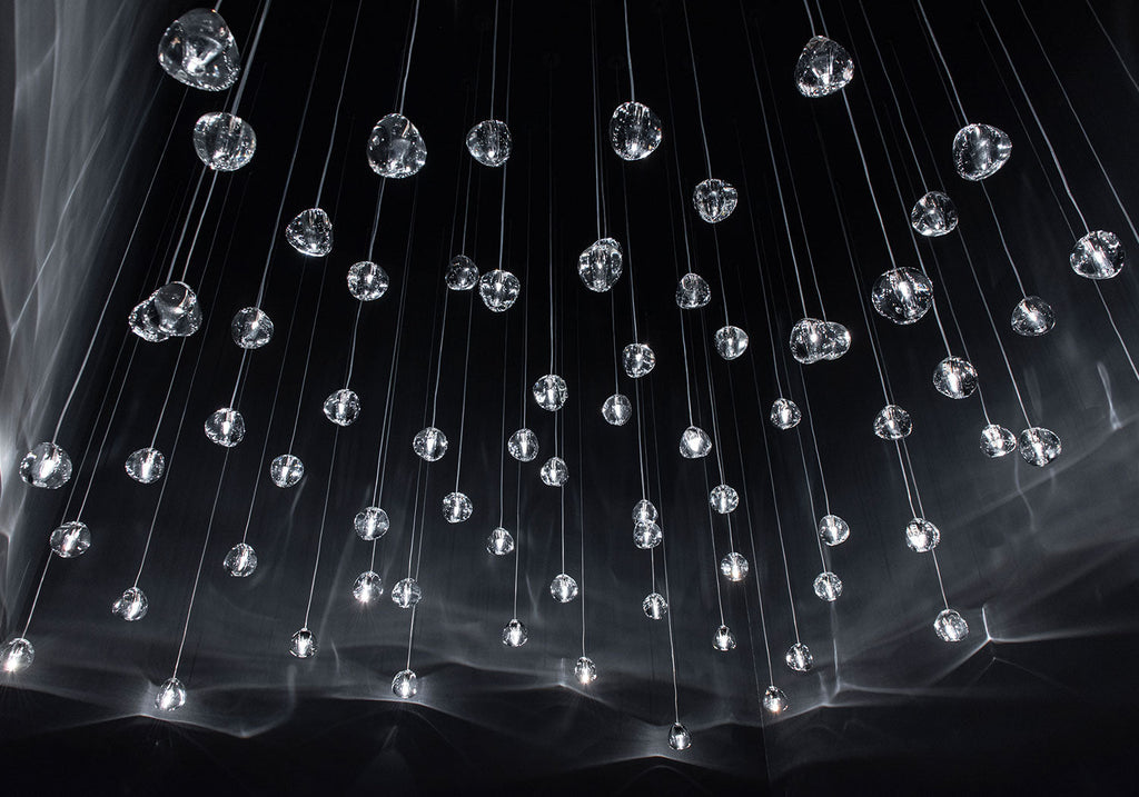 Looking up at a dark ceiling, clear glass heads of this Clérachic pendant hang on hundreds of thin wires with streaks of light across the glass.