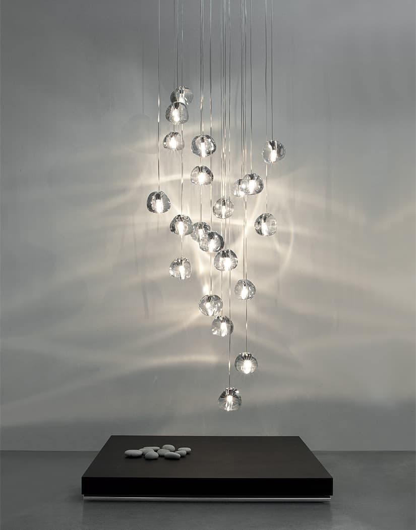 This Clérachic pendant hangs in a long cluster of clear glass heads in a grey room, with a low black plinth and pebbles below.